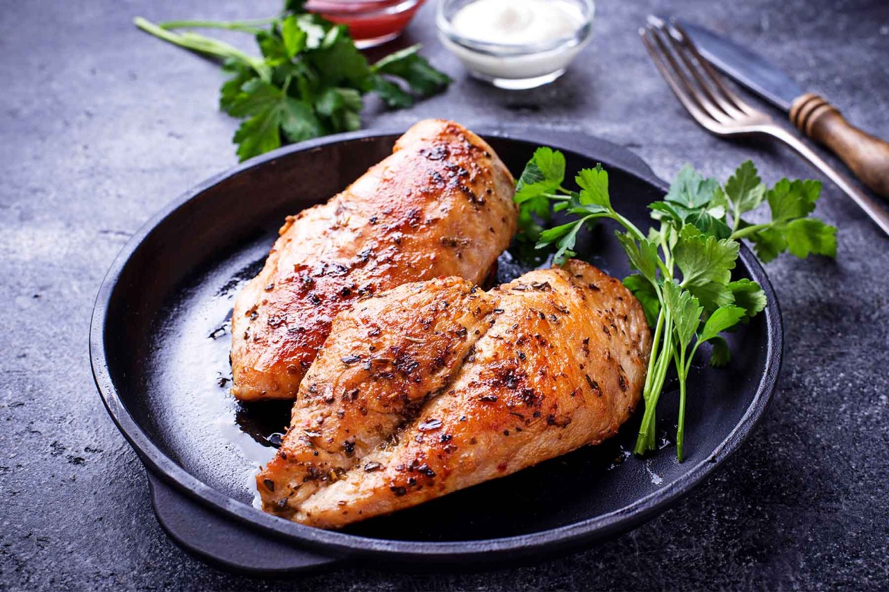 Two grilled chicken breasts with a golden-brown, herb-seasoned crust, served in a black cast iron skillet, garnished with fresh parsley, with forks and dipping sauces in the background.