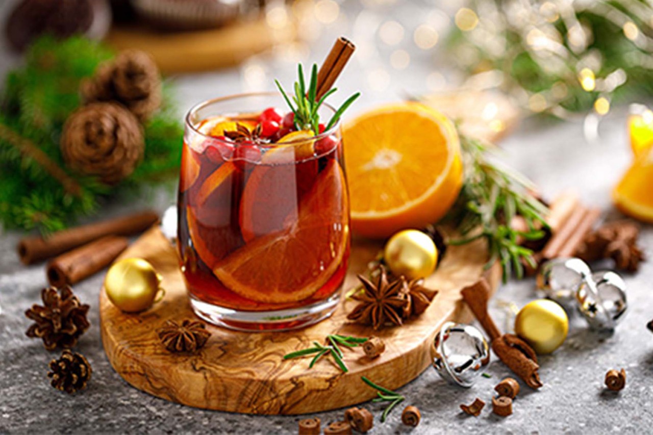 A festive drink in a glass, garnished with cinnamon sticks and fresh herbs, surrounded by orange slices, pine cones, and decorative ornaments on a wooden serving board, set against a cozy, seasonal backdrop.