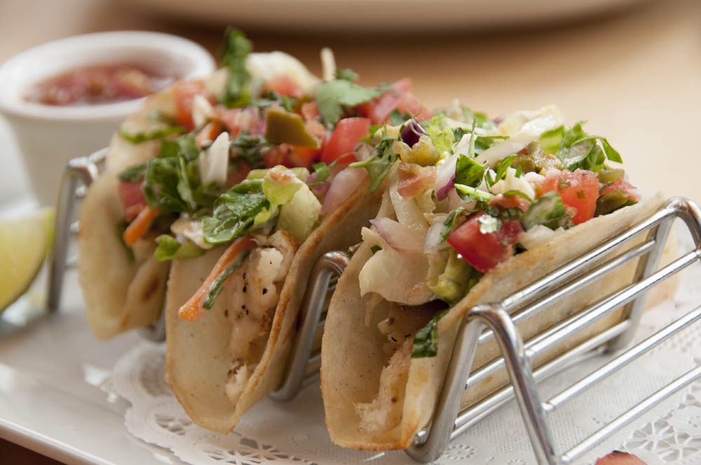 Three tacos filled with vegetables and garnished with herbs, served on a metal holder. A side of salsa is visible in the background.