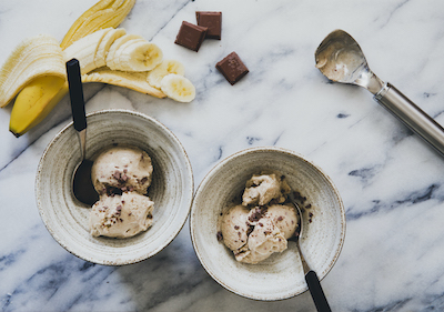 Two bowls of ice cream with chocolate chips, surrounded by banana slices, chocolate pieces, and an ice cream scoop on a marble surface.