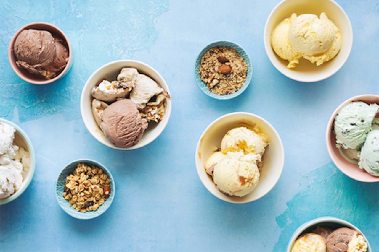 An assortment of ice cream scoops in various bowls, featuring flavors like chocolate, vanilla, and mint, arranged on a light blue surface.