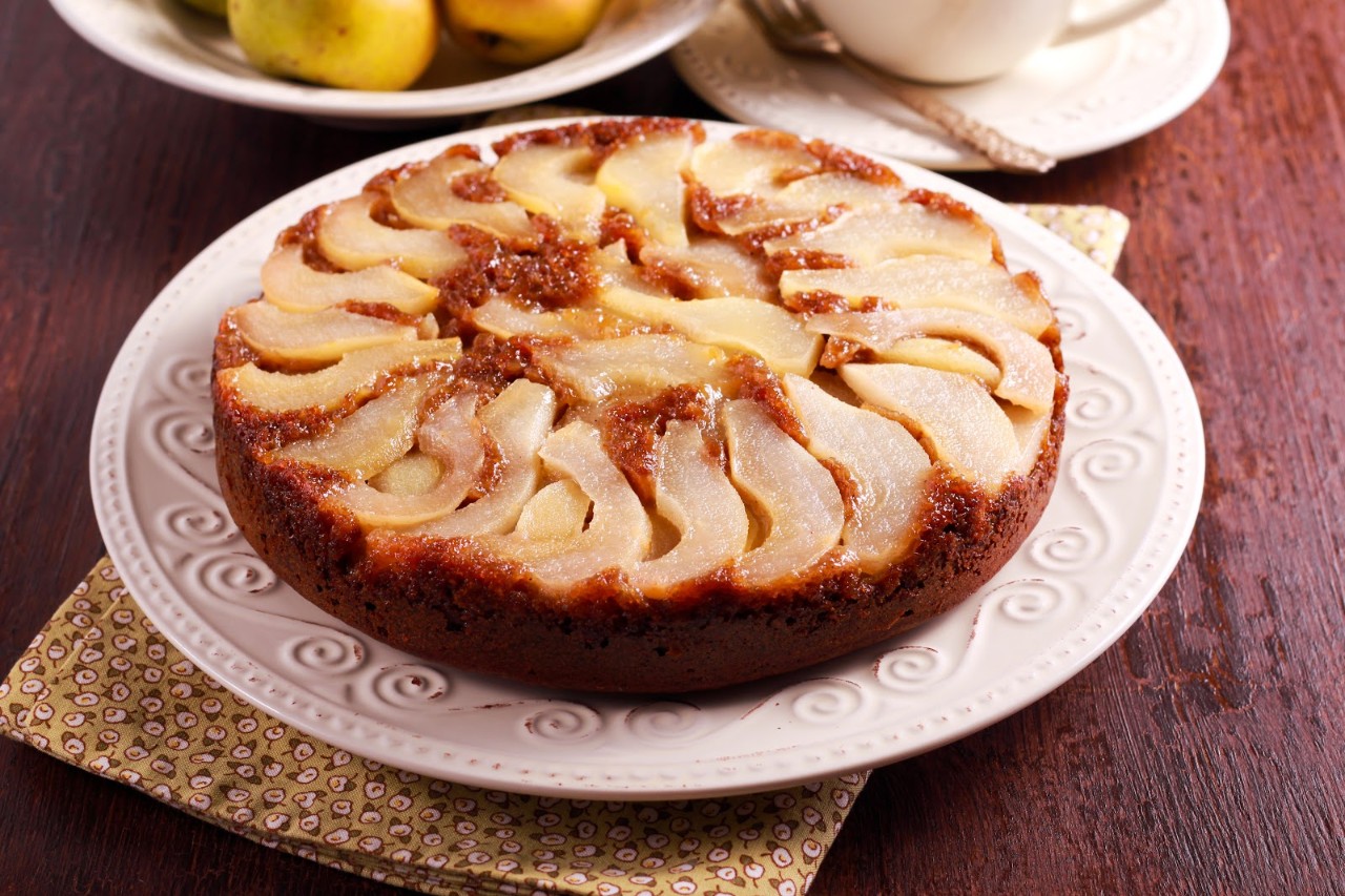 Round pear upside-down cake on a white plate, set on a patterned napkin, with a bowl of pears and a cup in the background.
