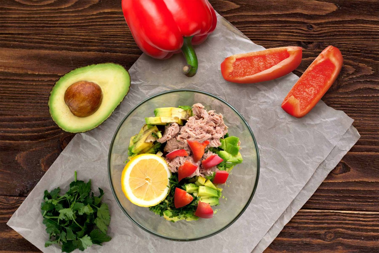 A fresh salad bowl with tuna, avocado slices, chopped lettuce, and strawberries, garnished with lemon, surrounded by red bell pepper halves and fresh cilantro on a wooden surface.