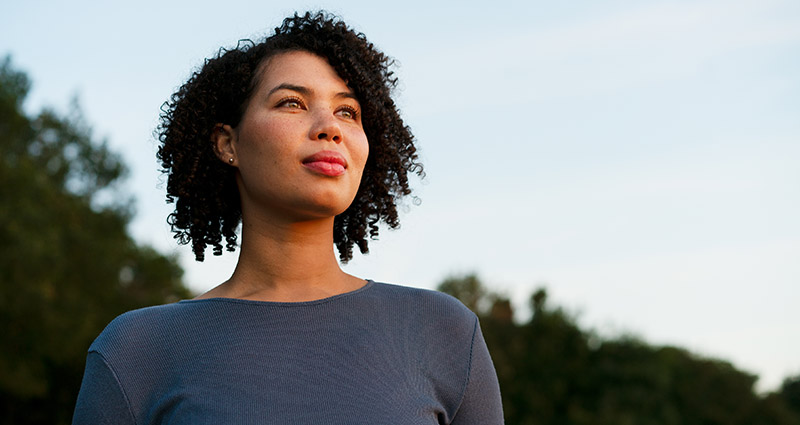 Woman with curly hair standing outdoors, looking confidently into the distance with trees and sky in the background.