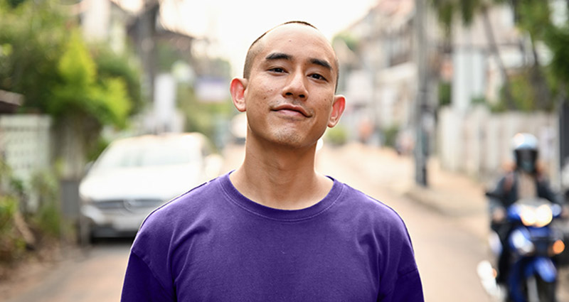 Confident young man with a shaved head wearing a purple shirt, standing on a city street with parked cars, trees, and a motorcyclist in the background.