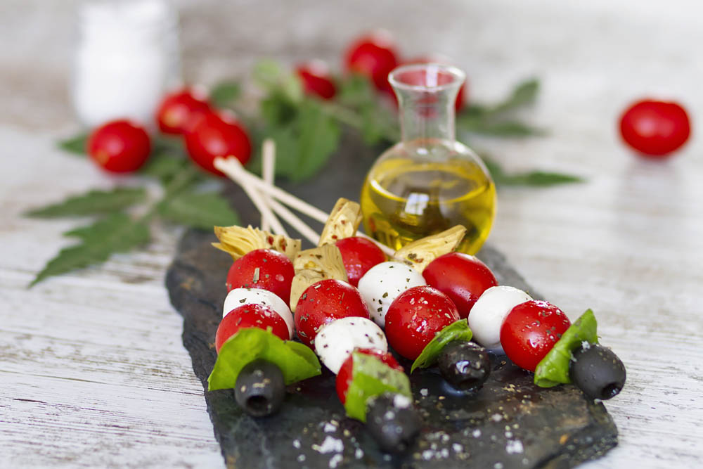Caprese skewers with cherry tomatoes, mozzarella, basil, and olives on slate; small bottle of olive oil nearby.