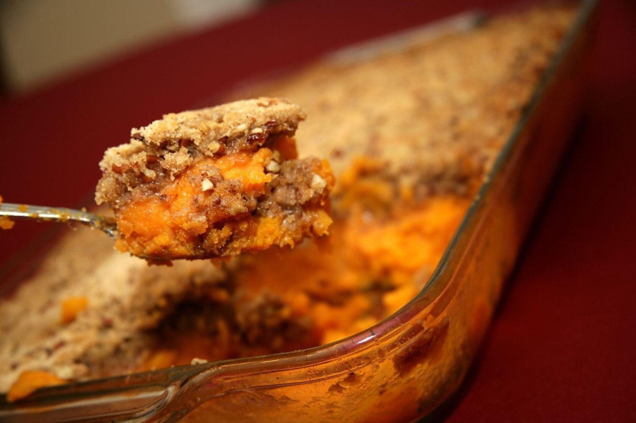 A serving of sweet potato casserole with a crumbly pecan topping being lifted from a glass baking dish.