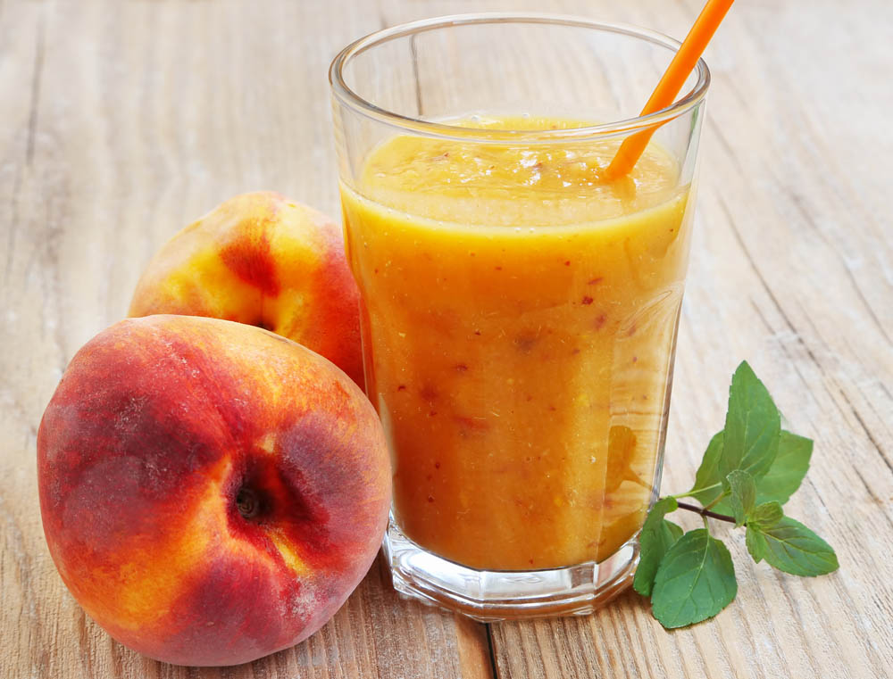 A glass of peach smoothie with an orange straw on a wooden table, next to two whole peaches and a small sprig of green leaves.