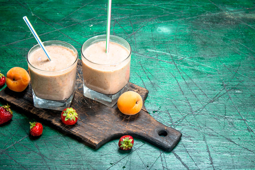 Two glasses of fruit smoothie on a wooden board with strawberries and apricots on a scratched green surface.