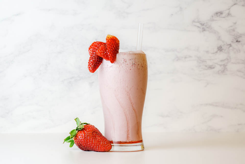 A glass of strawberry smoothie topped with strawberries, with a straw, placed on a white marble surface with more strawberries beside it.