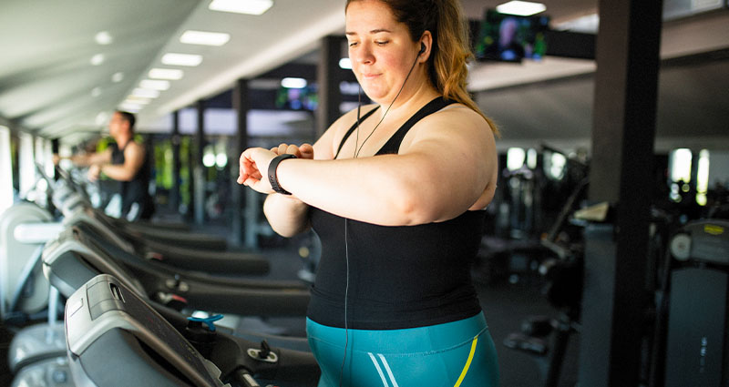 Focused woman using a treadmill in a modern gym, checking her smartwatch during a workout, surrounded by exercise equipment and large windows letting in natural light.