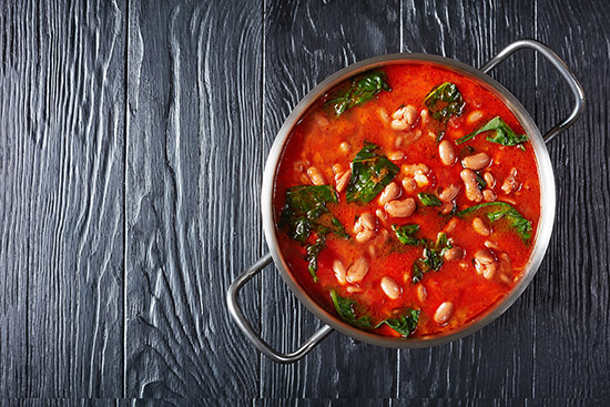 italian borlotti or cranberry beans soup with celery, vegetables, spinach, parmesan and tomatoes in a saucepan on a black wooden table, horizontal view from above, flatlay, copy space