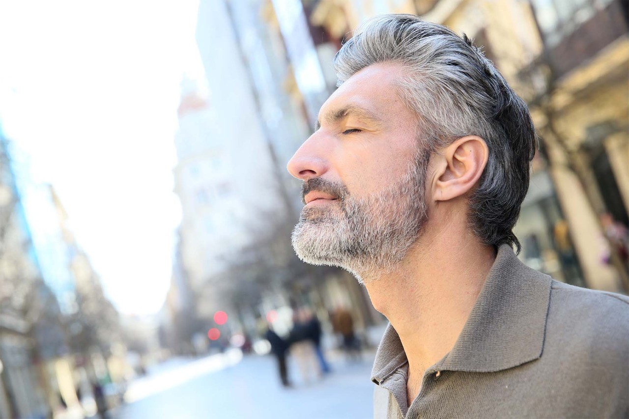 Middle-aged man with gray hair and beard standing outdoors in a city street, eyes closed and face tilted slightly upward, appearing to enjoy the fresh air and sunlight.