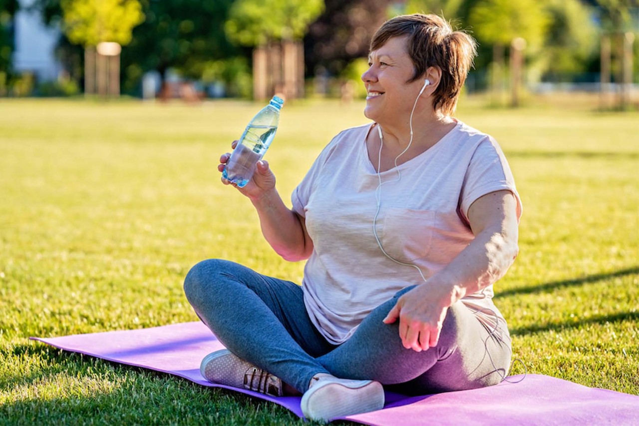 Middle-aged woman with short hair sitting cross-legged on a yoga mat in a park, holding a water bottle and smiling while listening to music through earphones, surrounded by green grass and trees.