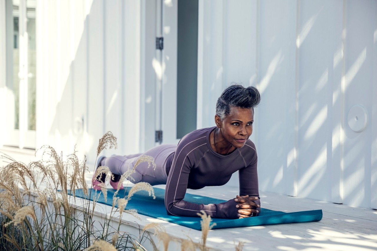 Elderly woman with short gray hair doing a plank exercise on a blue yoga mat outside, leaning on her forearms with a focused expression, surrounded by ornamental grasses against a white modern building.