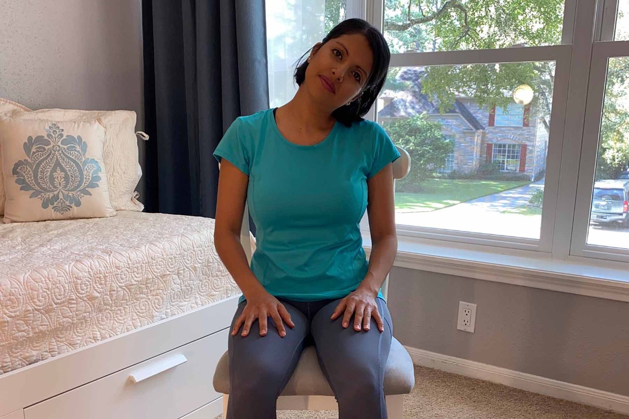 Woman with dark hair wearing a turquoise shirt and gray leggings sitting on a stool, tilting her head to the side as part of a neck stretch, hands resting on her thighs, with a window and a daybed with decorative pillows in the background