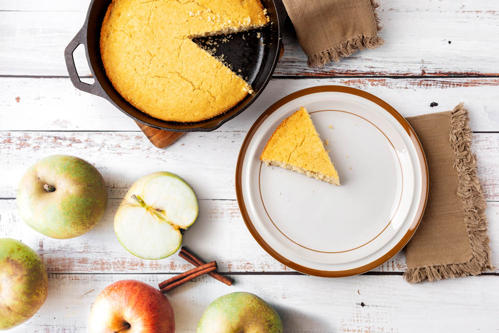 A cast iron skillet with cornbread missing a slice, a plate with the slice, apples, cinnamon sticks, and brown napkins on a wooden surface.
