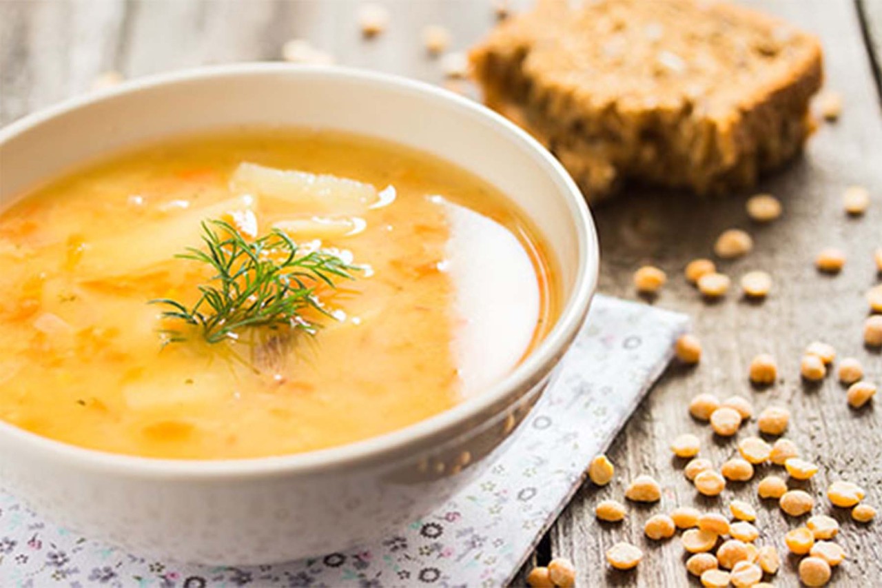 A bowl of creamy soup garnished with a sprig of dill, accompanied by a slice of brown bread and scattered peas on a rustic wooden surface.
