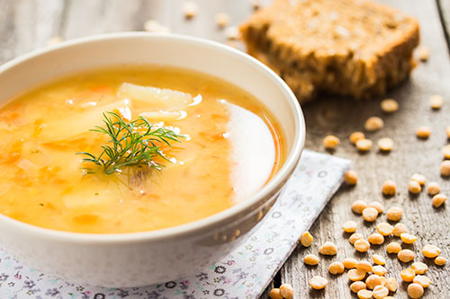 Bowl of yellow split pea soup garnished with dill, with a slice of bread and dried peas on a wooden table.