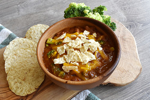 Bowl of chili topped with cheese and tortilla chips, served with broccoli and tortilla rounds on a wooden board.