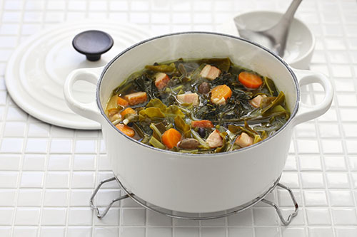 White pot containing soup with carrots, greens, and diced meat on a tiled surface, with a ladle and lid nearby.
