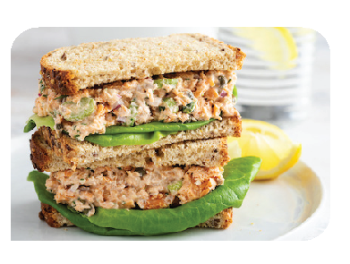 A stacked sandwich with tuna salad and lettuce on whole grain bread. A lemon wedge and a glass of water with a lemon slice are in the background.