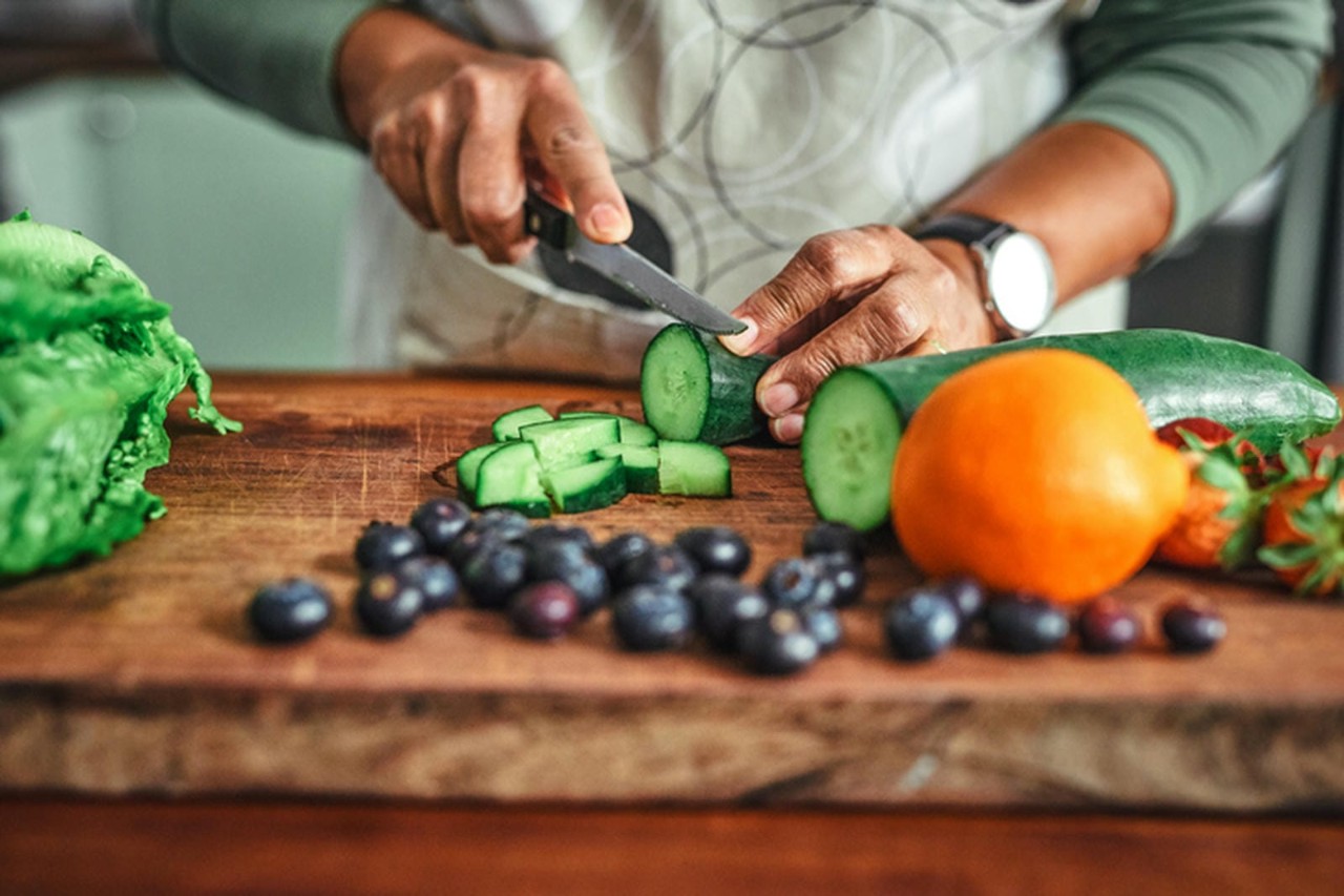 Hands slicing a cucumber on a wooden cutting board with lettuce, an orange, strawberries, and blueberries.