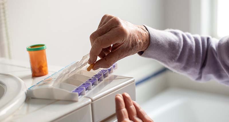 Close up of a person's hand taking pills out of a pill box that's sitting on a white bathroom sink counter
