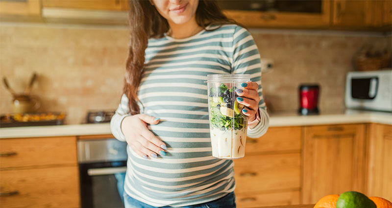 Pregnant woman holding a smoothie with spinach, blueberries, and banana.