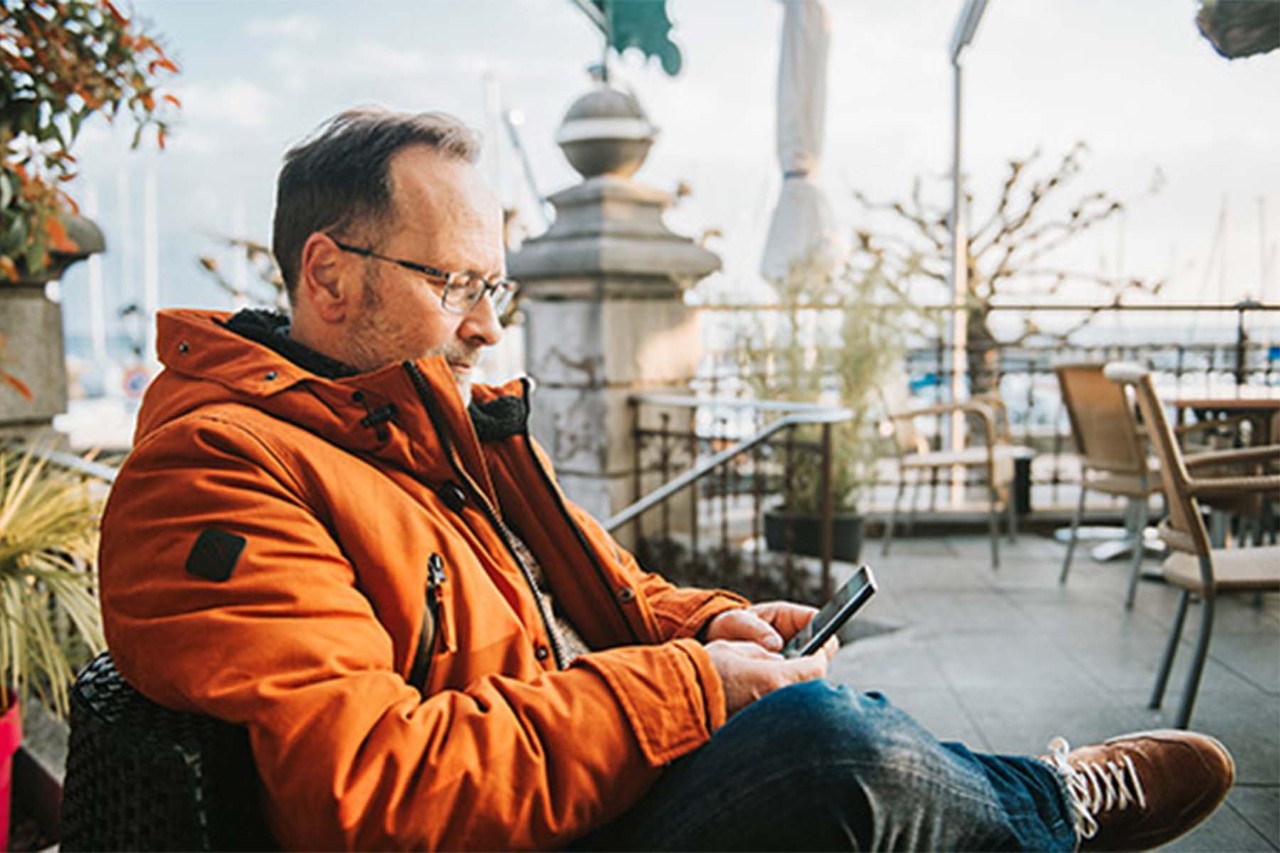 Man in an orange winter coat using his smartphone at an outdoor café with a marina in the background.
