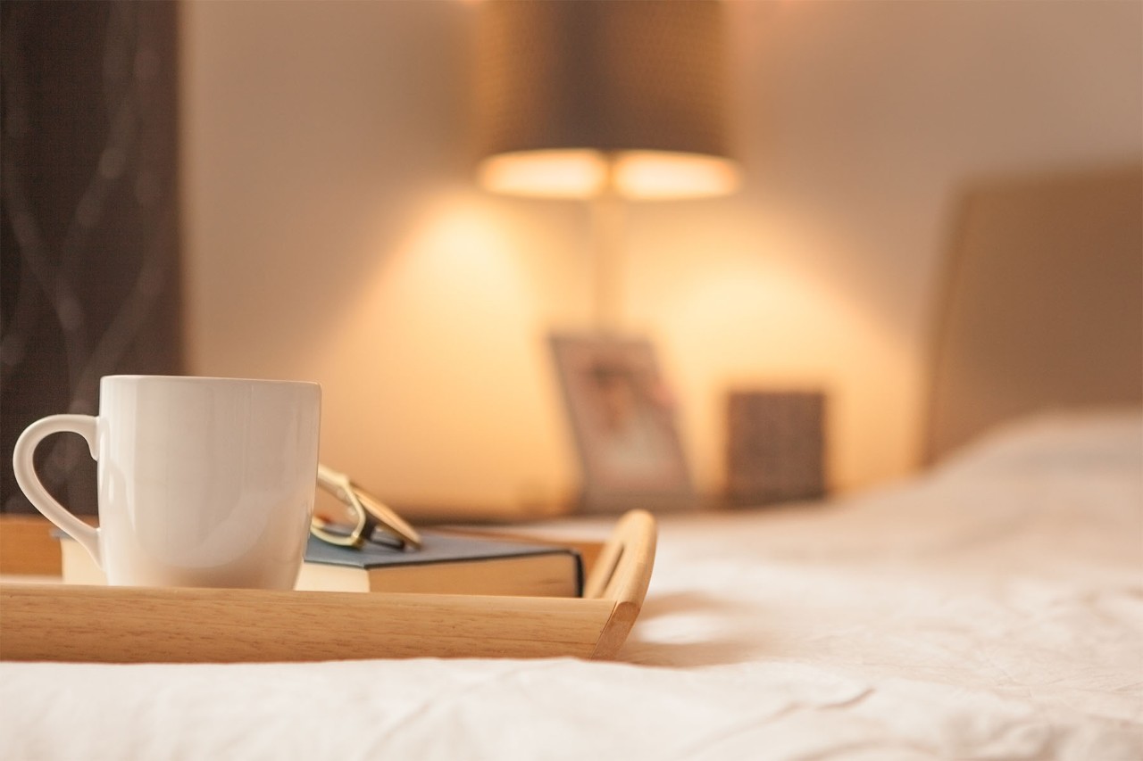White mug and book on a wooden tray atop a bed, with a warm-lit lamp in the background.