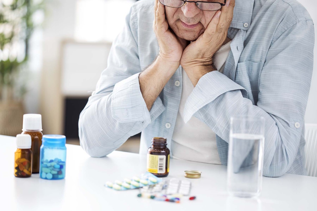 Older man with glasses holding his face in his hands, looking at various medication bottles and pills on a table.