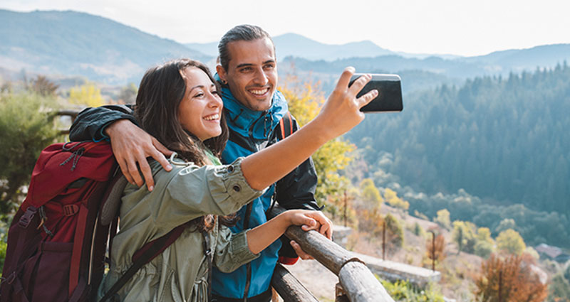 Smiling couple taking a selfie on a hiking trail with mountains and forests in the background.