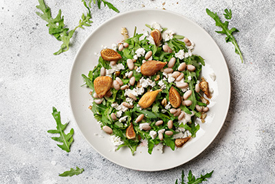 Plate of arugula salad with figs, white beans, and crumbled cheese on a textured surface.