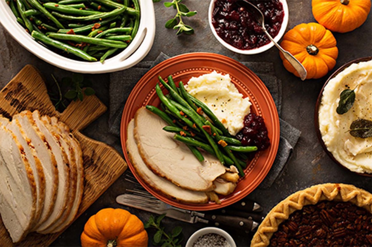 A festive Thanksgiving spread featuring sliced turkey, green beans, cranberry sauce, mini pumpkins, and a pumpkin pie, all arranged on a wooden table.
