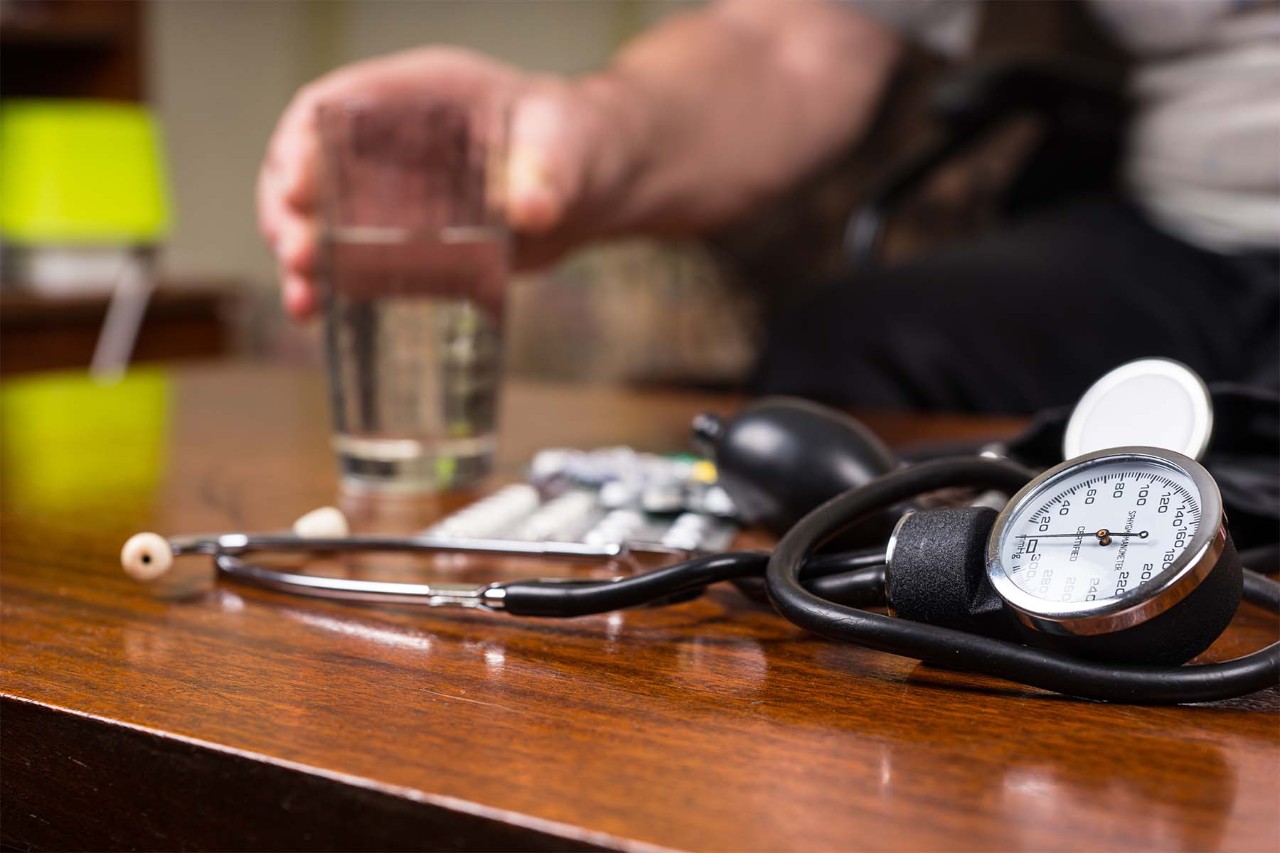 Blood pressure monitor and stethoscope on a wooden table, with a person reaching for a glass of water in the background.