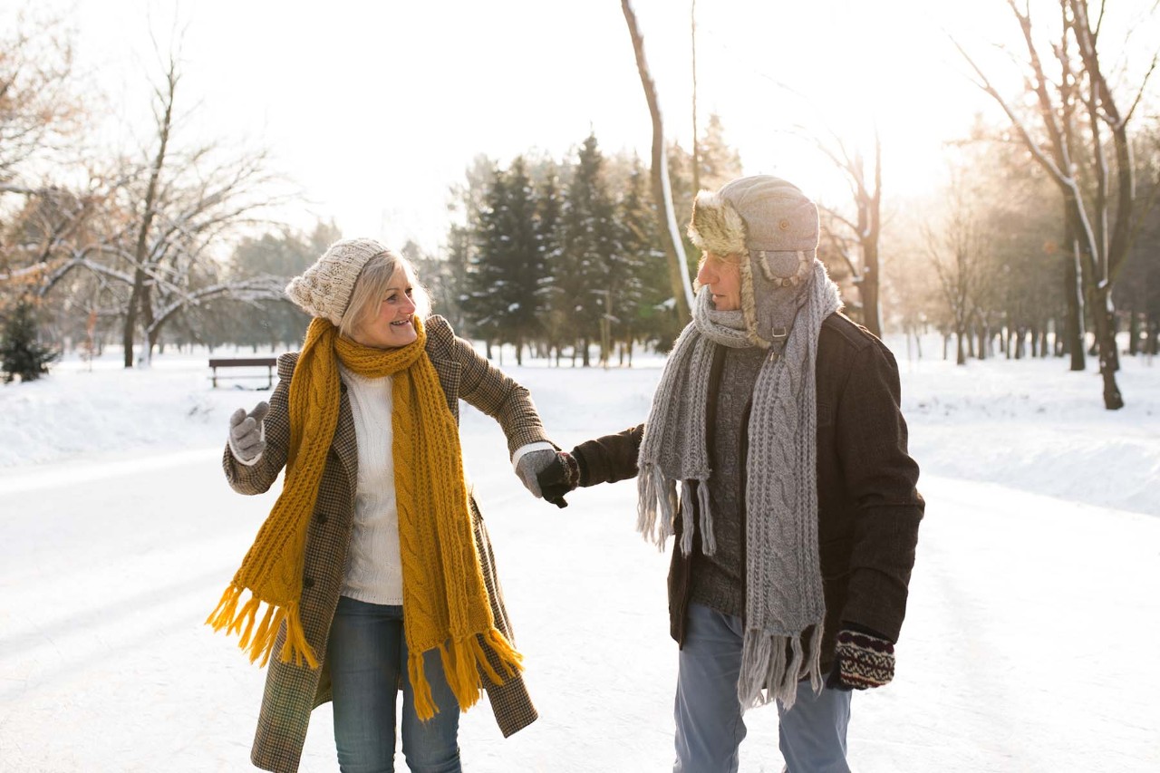 Older couple walking hand in hand on a snowy path, bundled up in winter coats, scarves, and hats, with the sun shining through the trees.