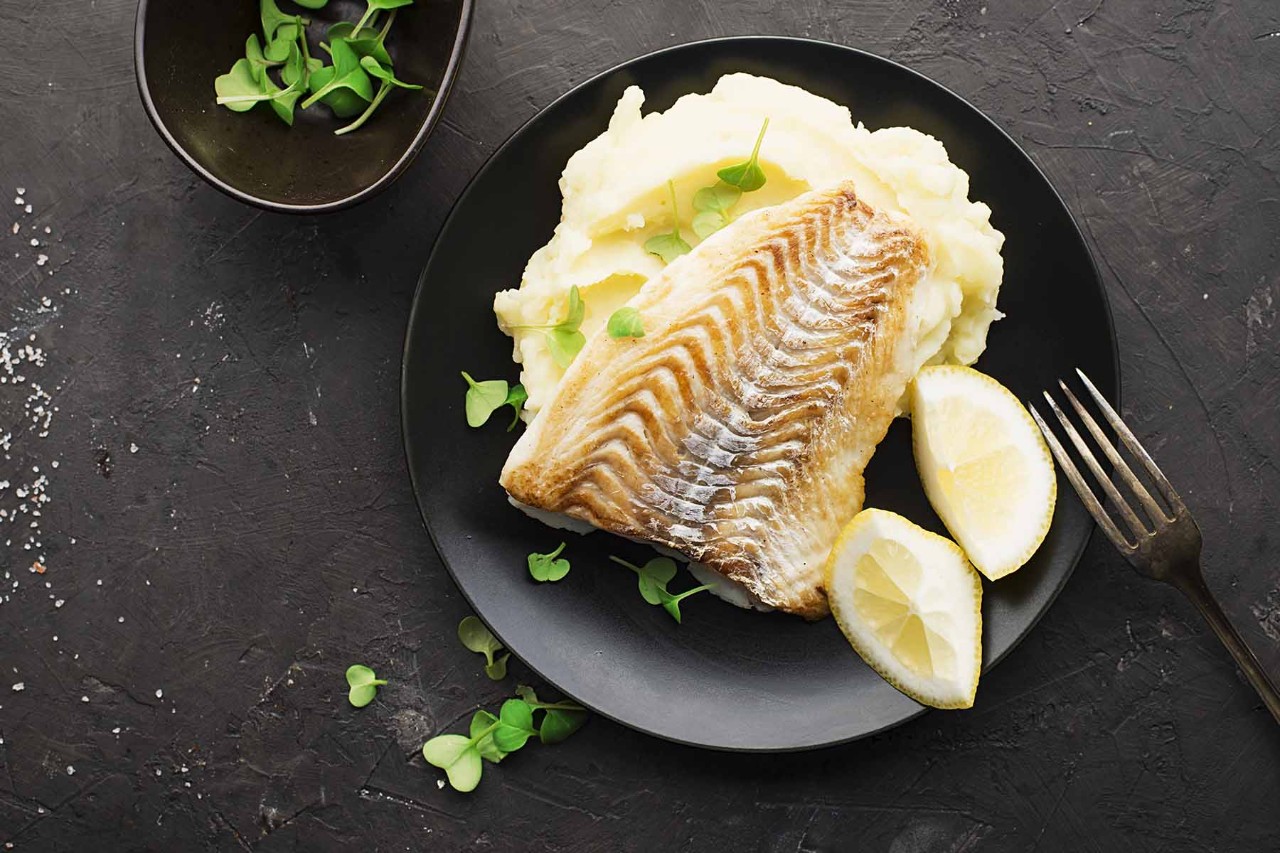 A plate featuring a portion of grilled fish with lemon wedges, served alongside creamy mashed potatoes and garnished with fresh herbs, on a dark textured surface.