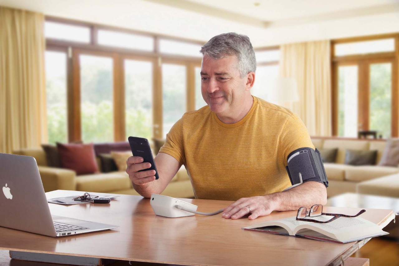 Middle-aged man using a blood pressure monitor on his arm while looking at his smartphone, seated at a table with a laptop and open book.