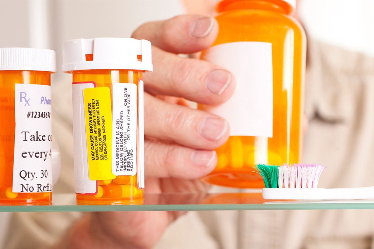 Hand reaching for an orange prescription bottle among other medication containers on a glass shelf, with a toothbrush nearb
