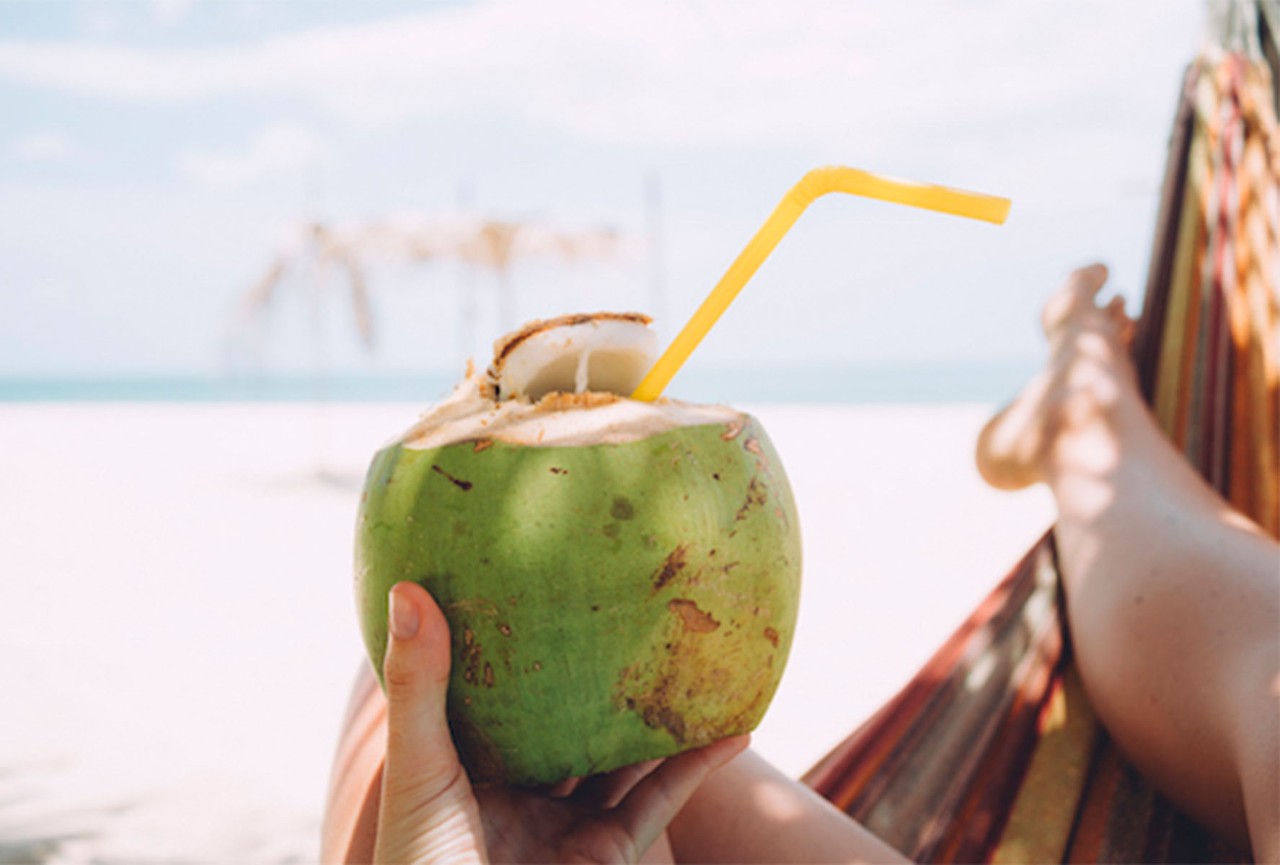 Person relaxing in a hammock on a sandy beach, holding a green coconut with a yellow straw, with clear blue skies and distant palm trees in the background.