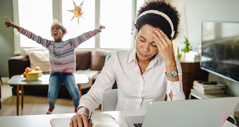 Woman working on a laptop with her hand on her head, while a child plays energetically in the background.