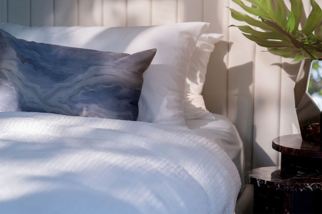 A cozy bedroom corner featuring a neatly made bed with white linens and a decorative pillow, alongside a small side table with a plant, creating a serene atmosphere.