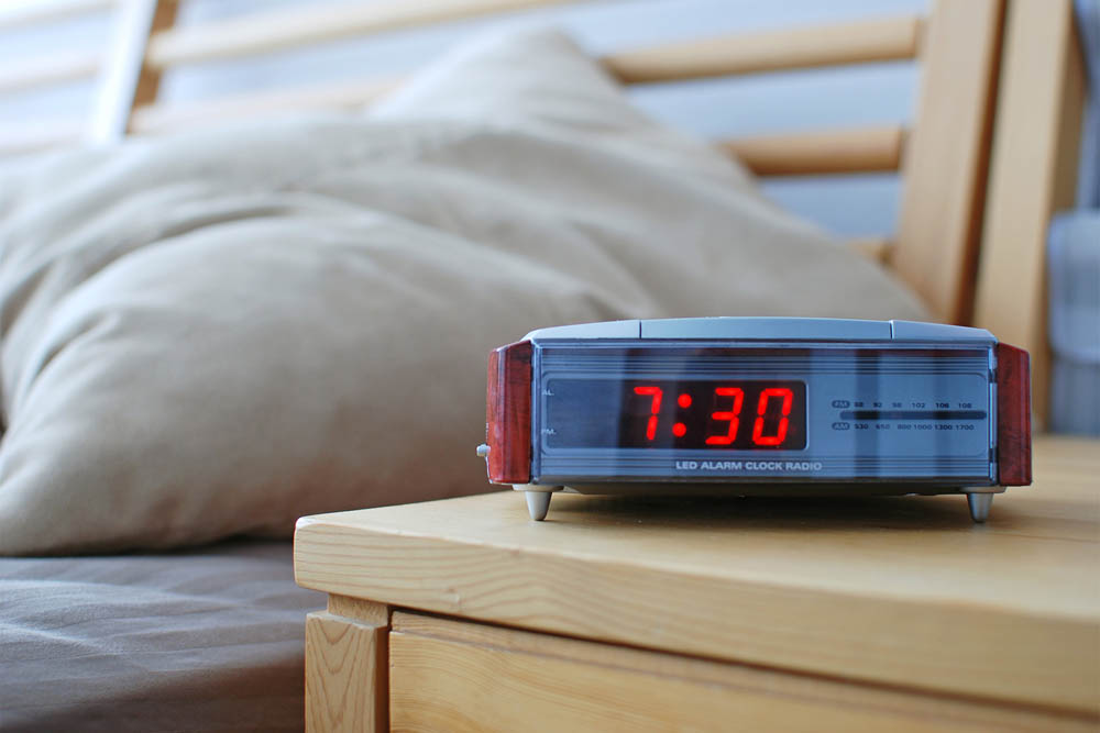 A digital alarm clock displaying the time 7:30 on a bedside table, with soft pillows in the background, creating a cozy bedroom atmosphere.