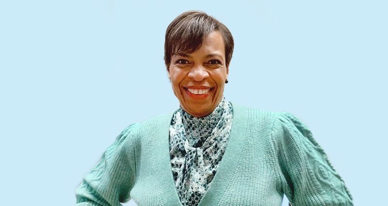 Smiling woman wearing a green sweater and patterned blouse, standing against a light blue background.