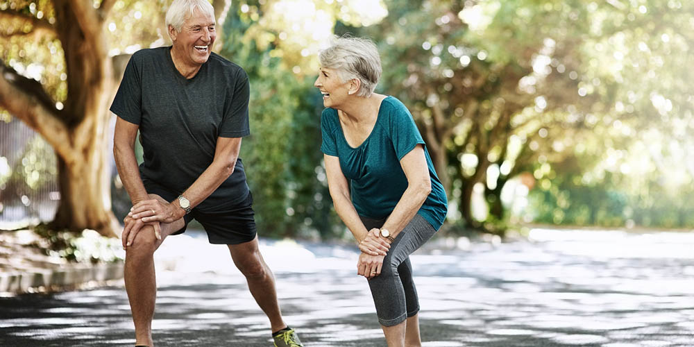 An elderly couple stretches outdoors on a sunny day, wearing comfortable exercise clothing, with trees in the background.