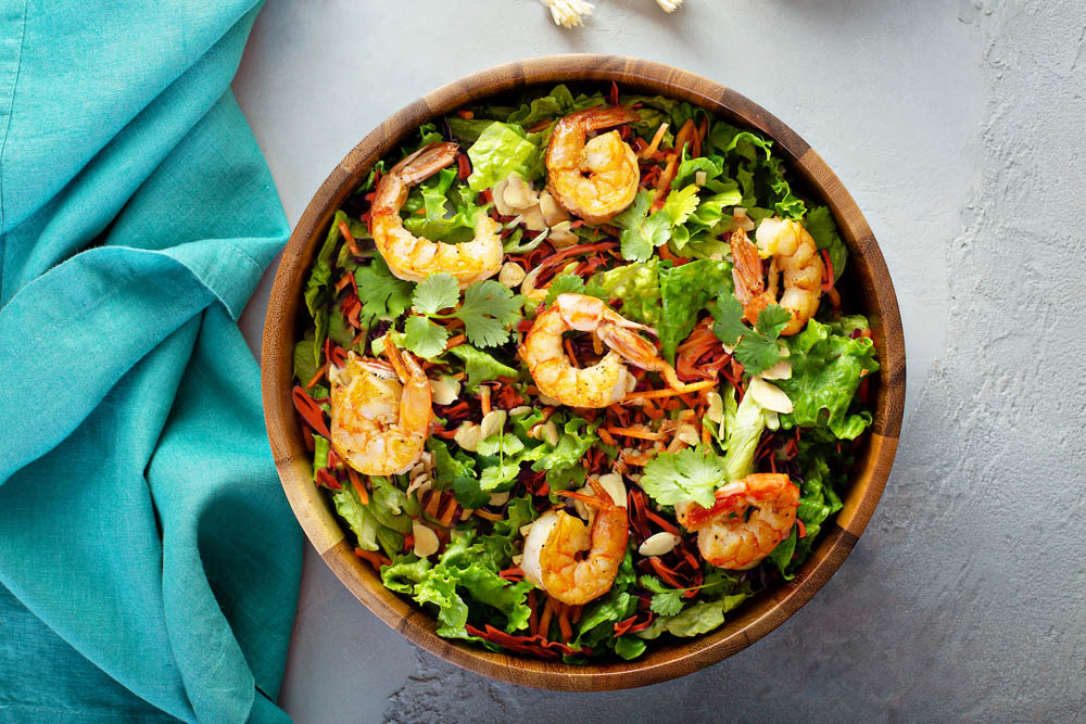 A wooden bowl filled with shrimp salad, including lettuce, carrots, and almonds, on a gray surface next to a blue cloth.