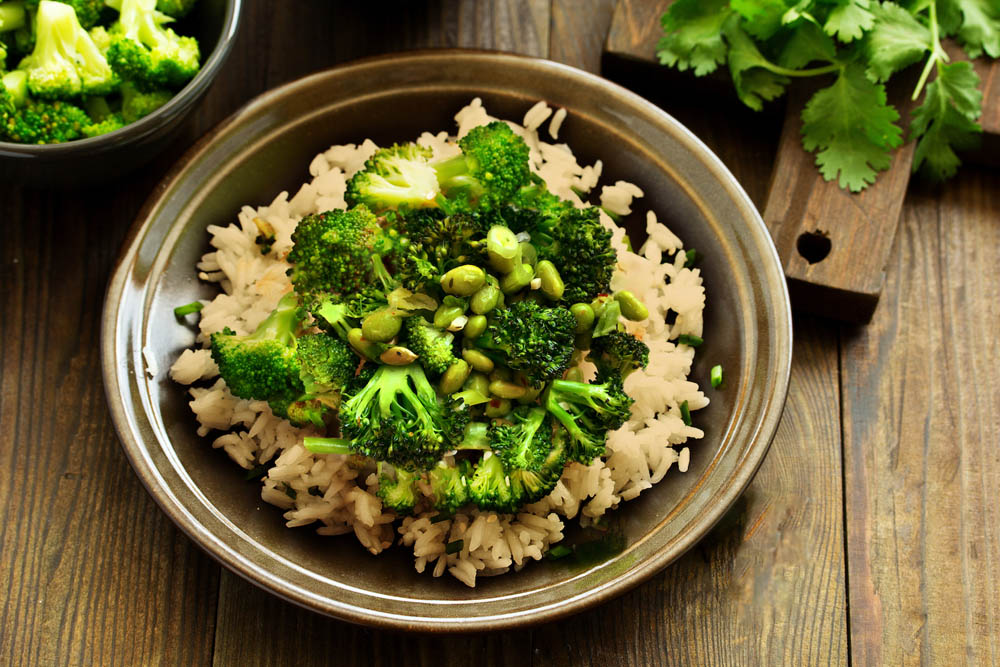 A plate of rice topped with broccoli and edamame, garnished with herbs, on a wooden table.