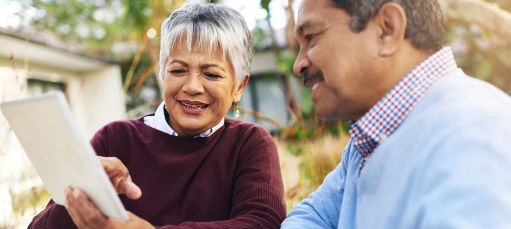 Two older adults sitting together outdoors, one holding a tablet and both smiling as they engage with each other in conversation, surrounded by greenery.