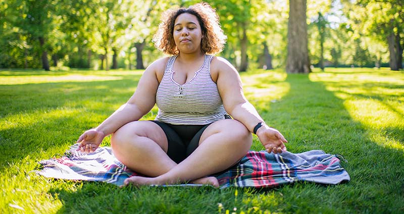A woman sits cross-legged on a blanket in a park, meditating with her eyes closed, surrounded by lush green trees and sunlight.